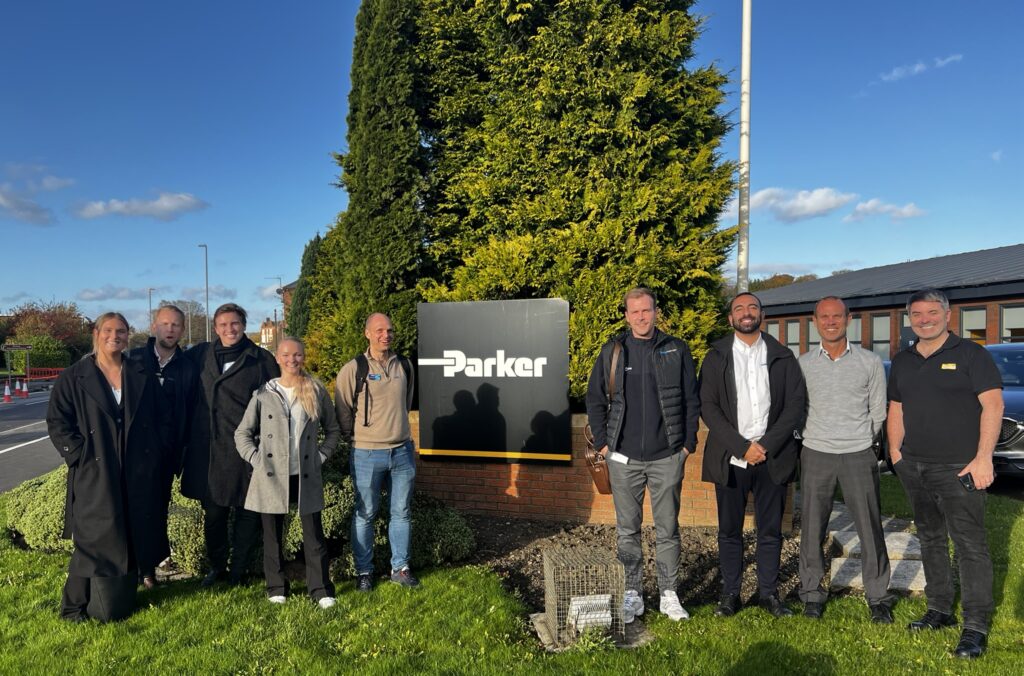Udendørs gruppebillede foran Parker Hannifin-skiltet med repræsentanter fra Colly Flowtech og Parker Hannifin under et fælles besøg. Solrig dag med blå himmel, grønt græs og smilende deltagere.
